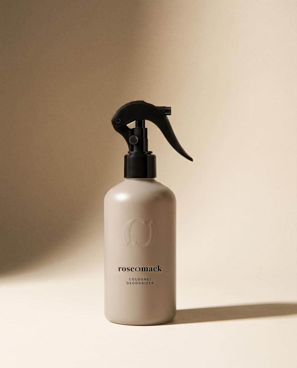 roseomack Cologne in a greige trigger-spray bottle, a soft raking shadow across the warm beige wall behind