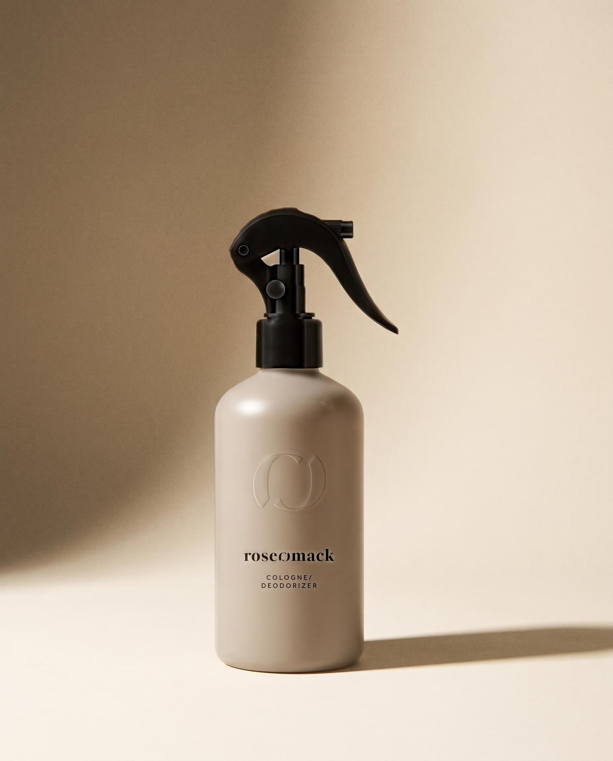 roseomack Cologne in a greige trigger-spray bottle, a soft raking shadow across the warm beige wall behind